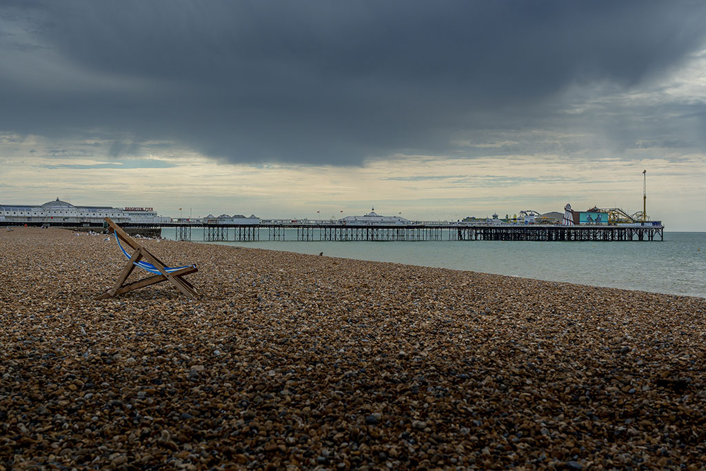 A moody sky overshadows a shingle beach on which a lone empty deckchair stands. A pier with funfair is in the middle distance.