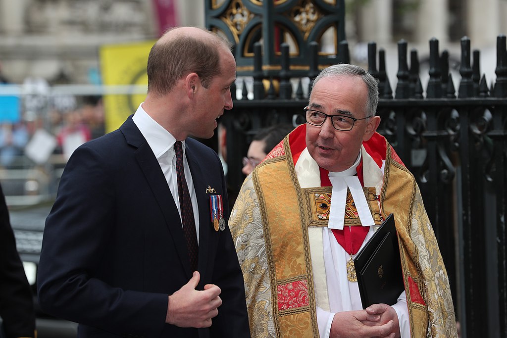 A young man wearing a dark suit talks to a minister wearing regalia.