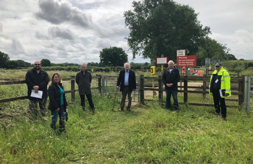 A group of people stand in a field by a fence and a railway footcrossing.