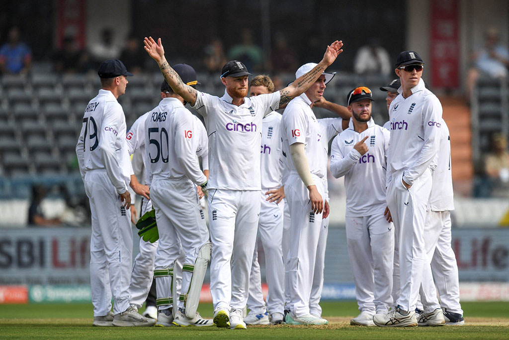 A gaggle of cricket players, dressed in whites, stand on the field. One raises there arm