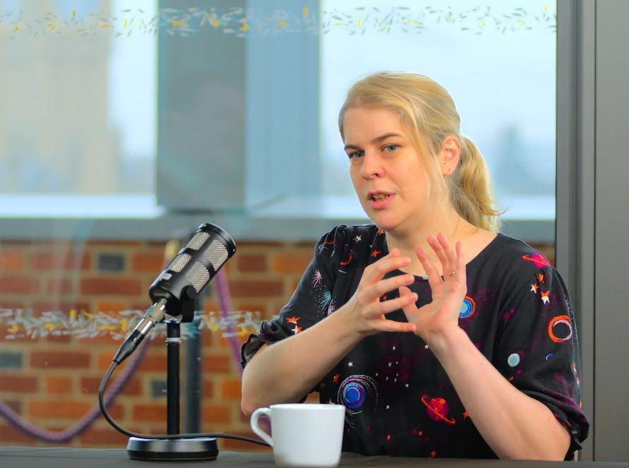 a woman sits at a table, with a mic in front of her, talking and holding her hands together in front of her.