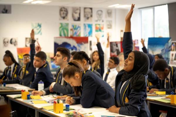 school pupils sit at desk, some with a hand raised.