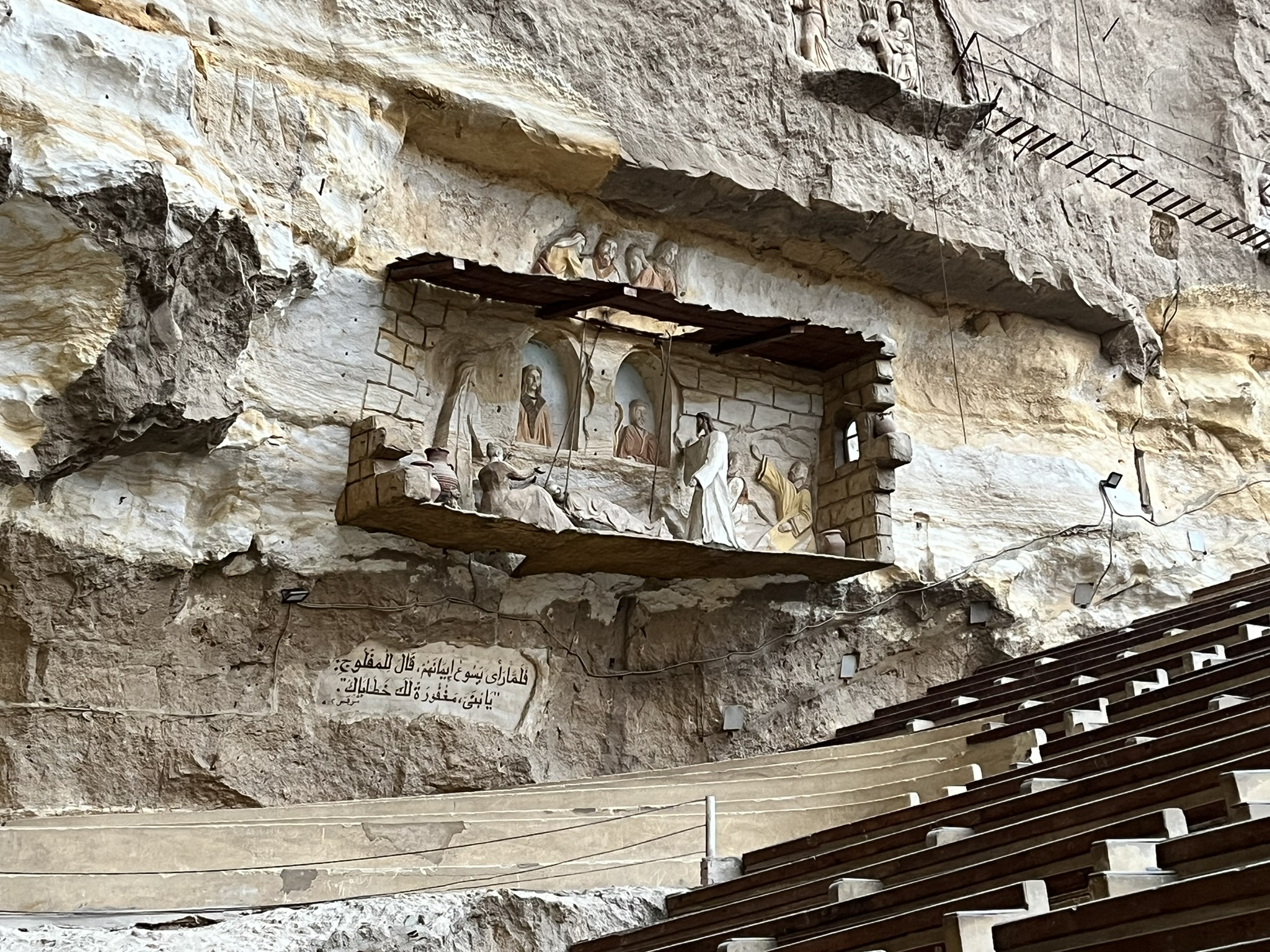 A bible scene is carved into the wall of a cave, below which is seating.