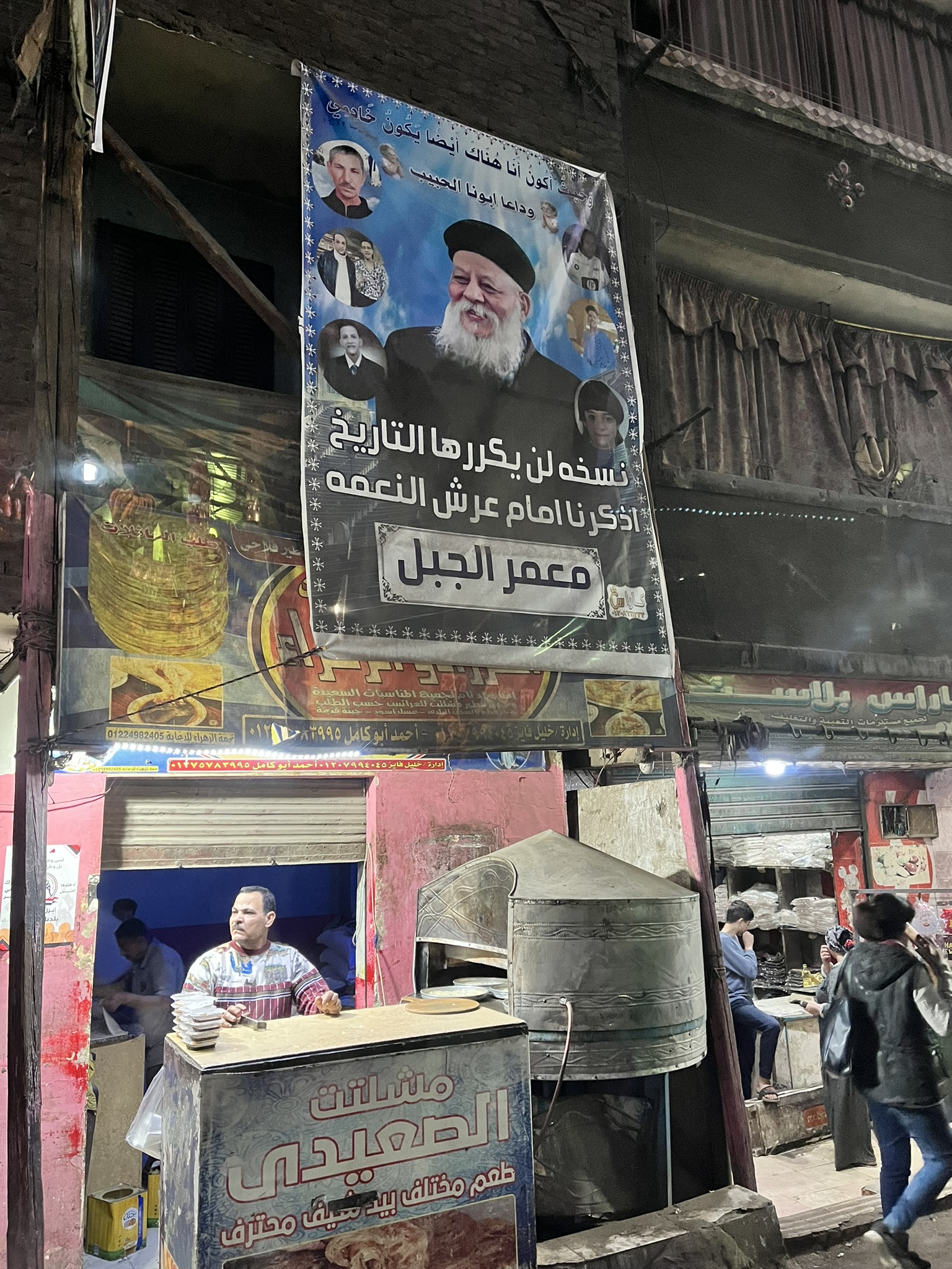 A street seller stands at a booth below a poster of a priest.