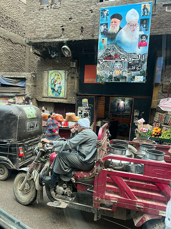 A motor-trike passes along a narrow street past a poster of a priest and a pope.