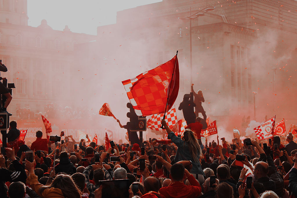 Silouhetted by red flare smoke, celebrating footballs wave red flags.