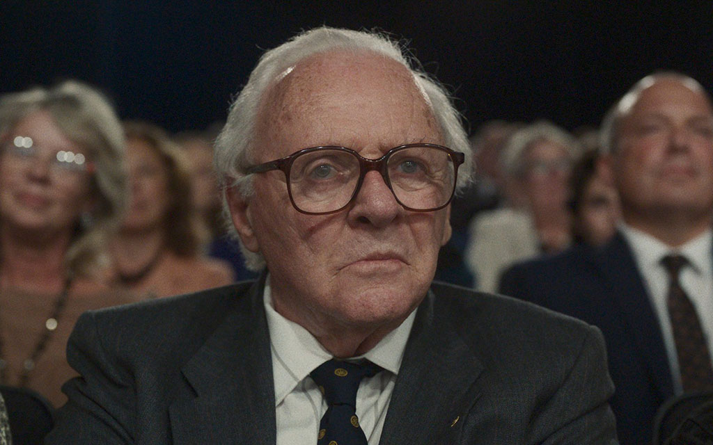 An old man wearing a suit and tie sits in a TV audience as people stand around him.