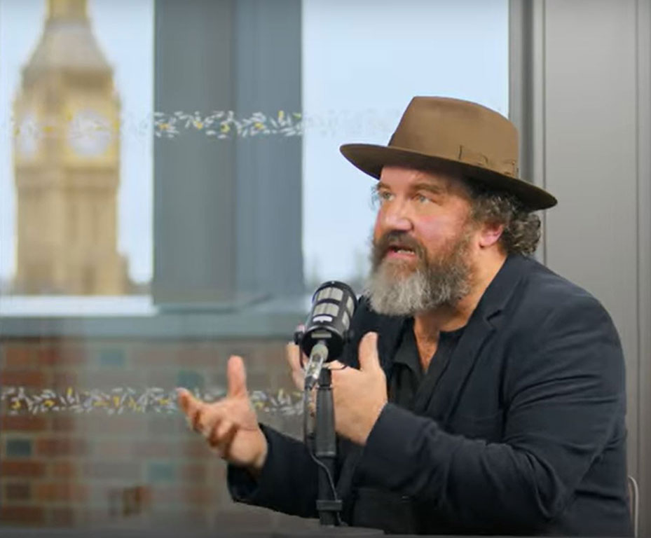 A man wearing a hat sits at a table talking and raises both hands in front of himself to gesture