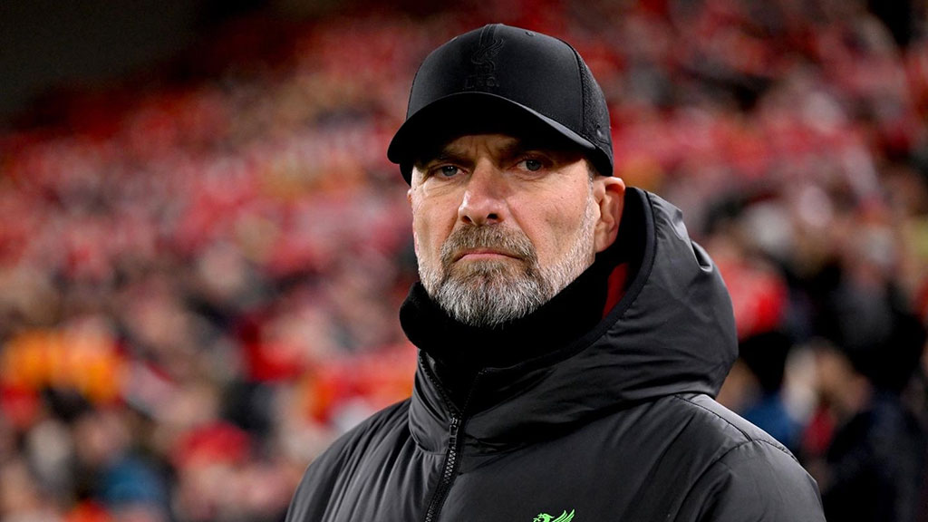 A football manager stands on the touchline and stares hard, dressed in a black coat and hat.