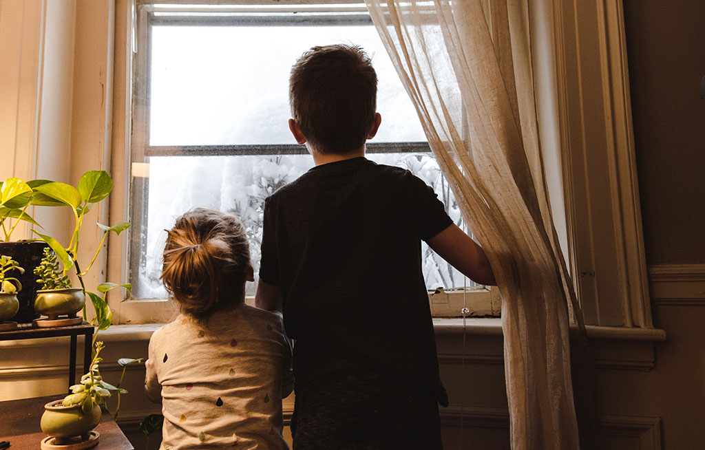 Two small chidlren push a curtain aside to stare out the window at snow.