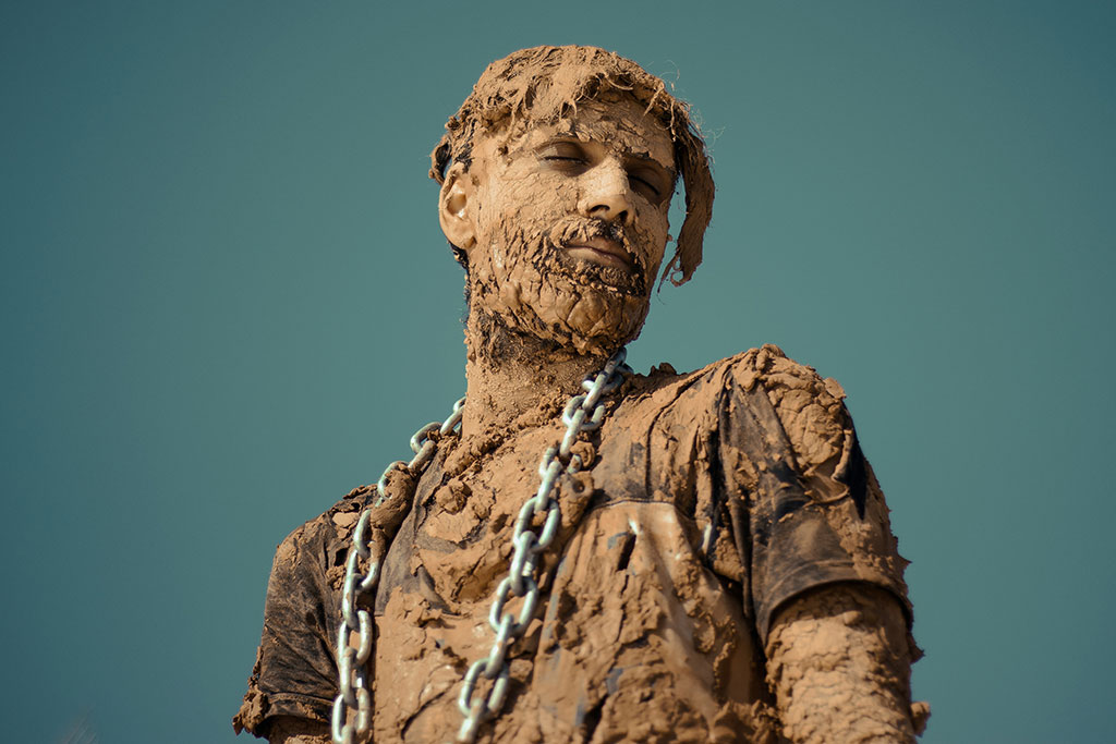 A man covered in dried and caked mud stands and looks to the side, a steel chain is draped from his shoulders.