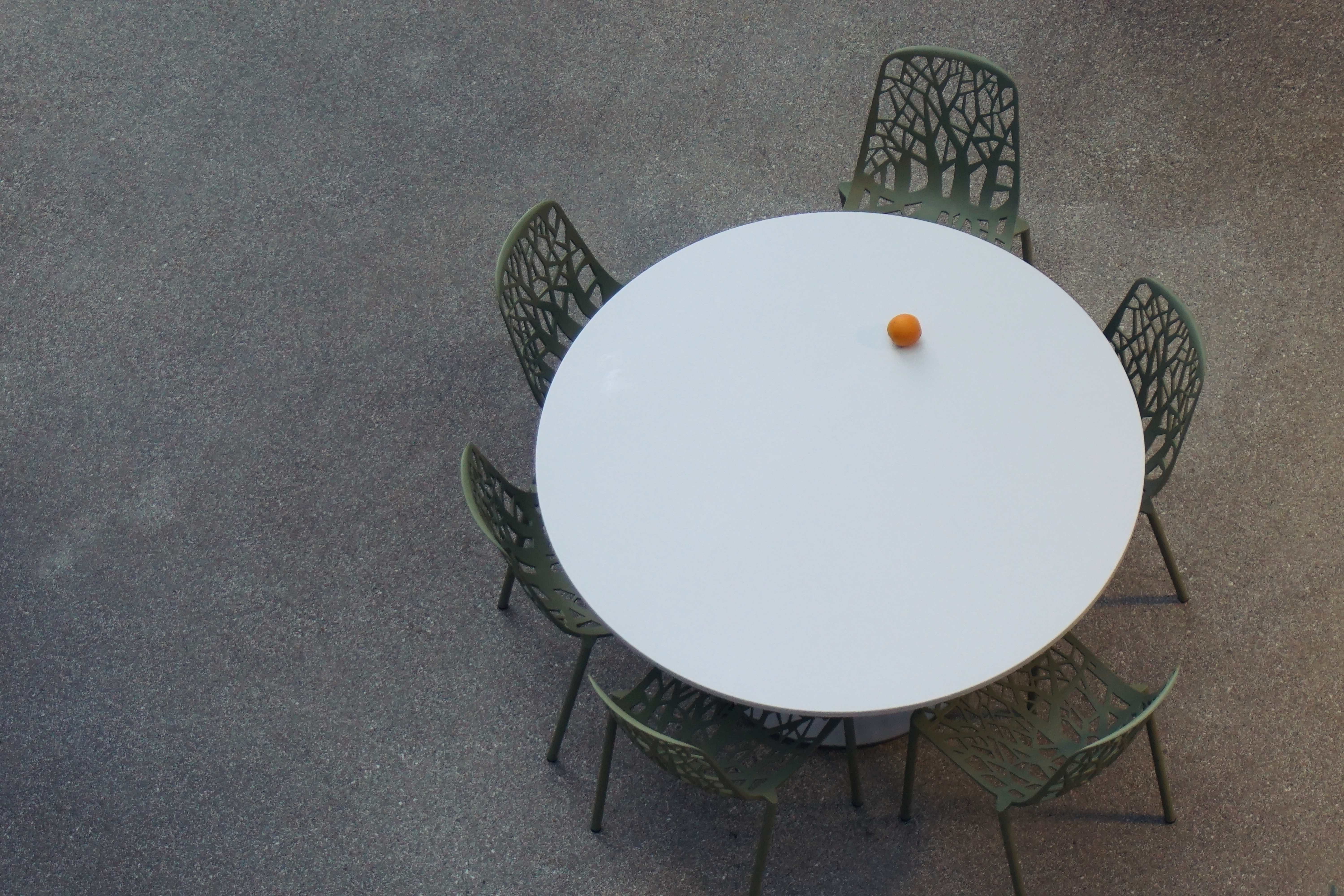 a round table with empty chairs is seen from above. An orange is the only item on it.