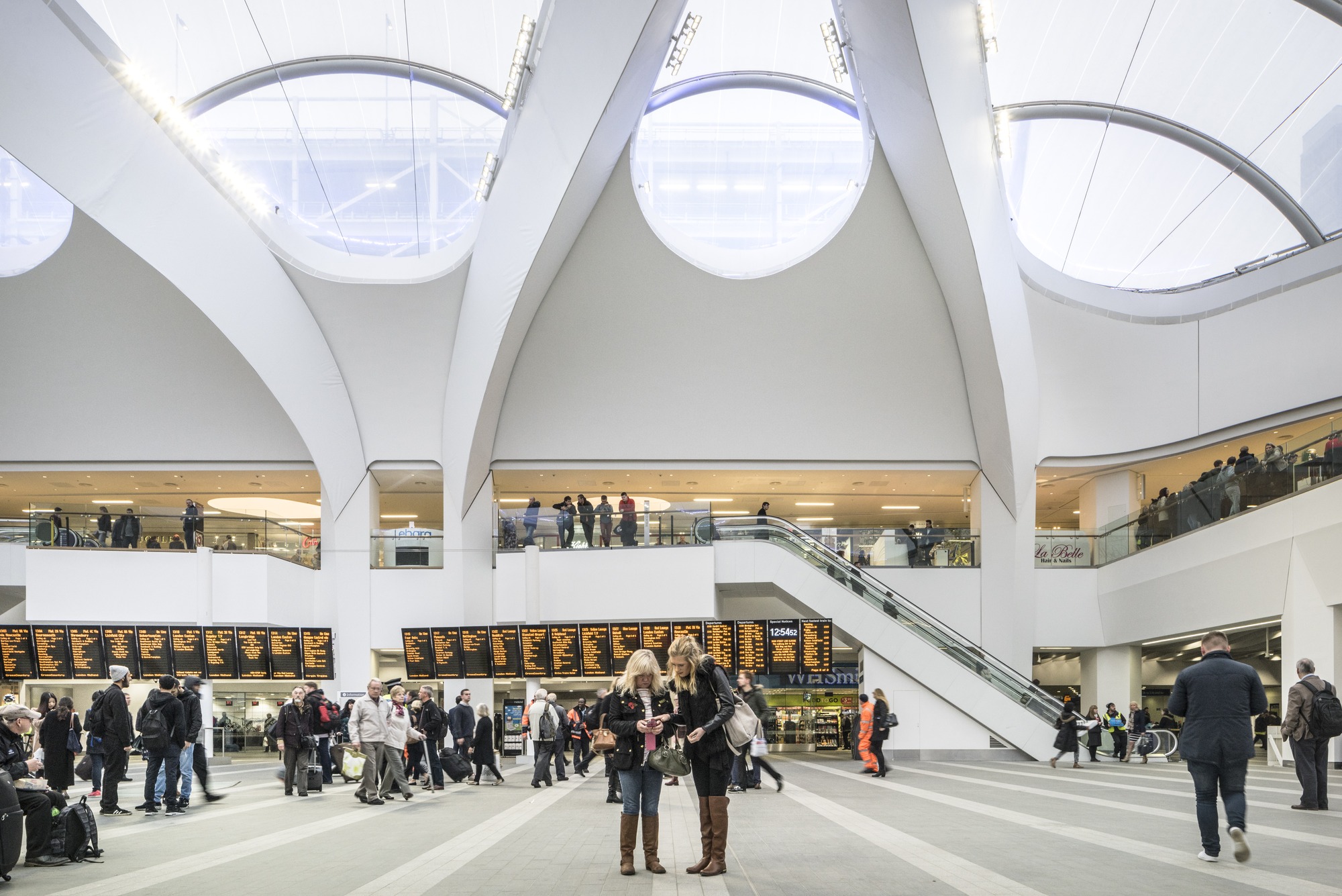 A airy and light station concourse in which people stand and look at a long set of travel information screen.
