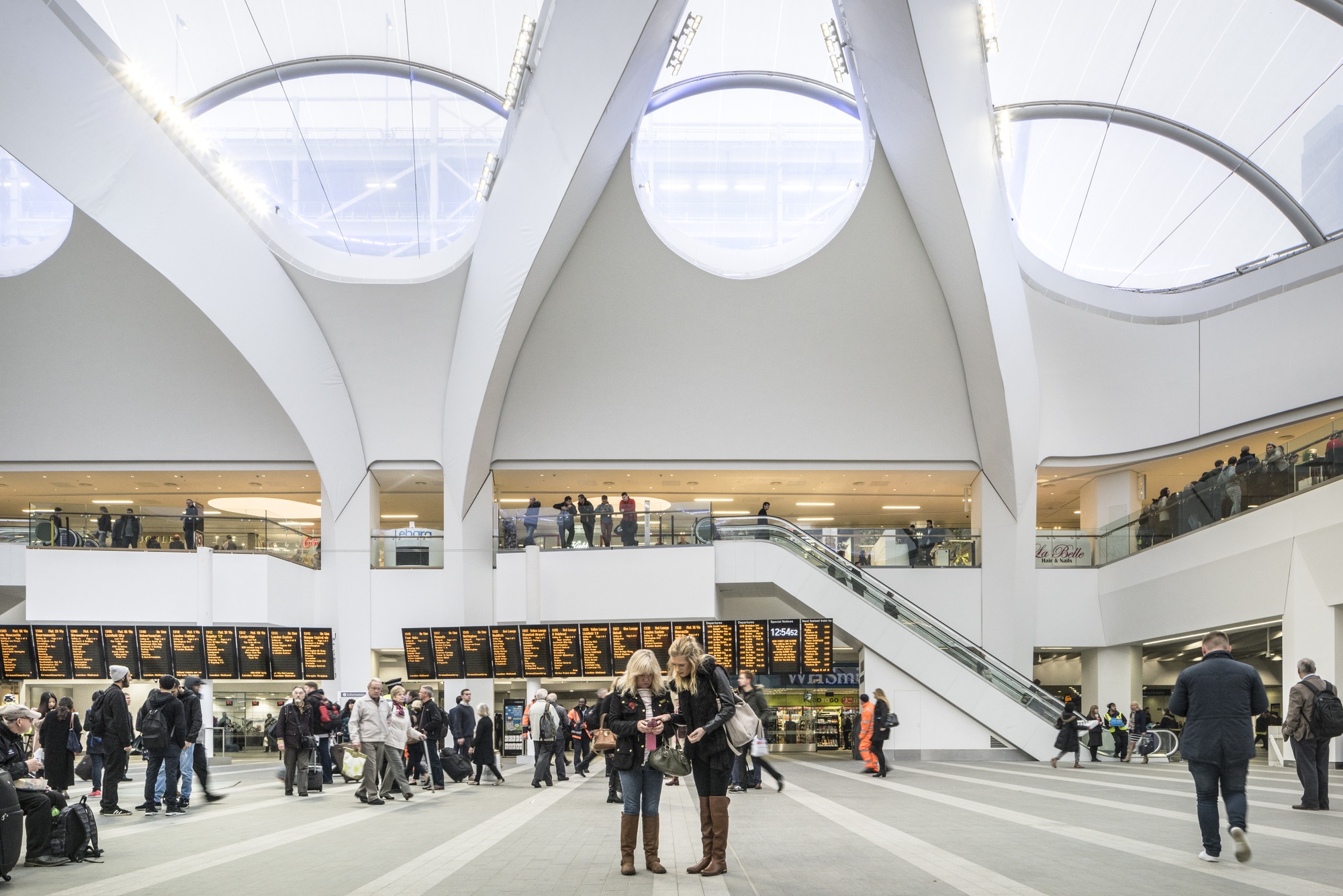 A airy and light station concourse in which people stand and look at a long set of travel information screen.