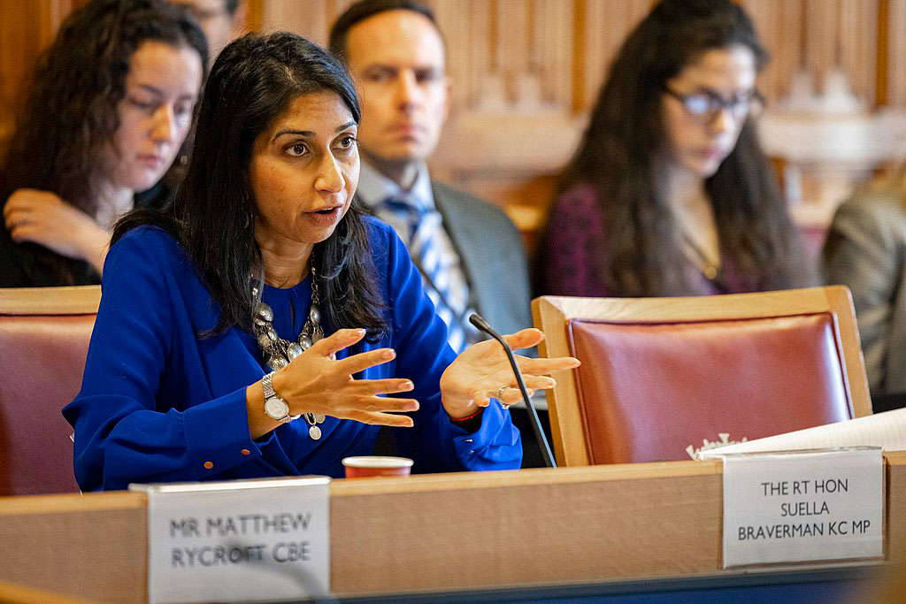 A woman dressed in a blue suit sits at a table talking and gesticulating with her hands.