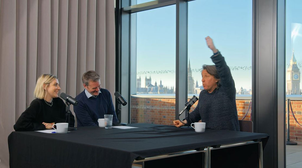 A sitting man raises one hand above his head to emphasise a point, while two other people looking on are laughing.