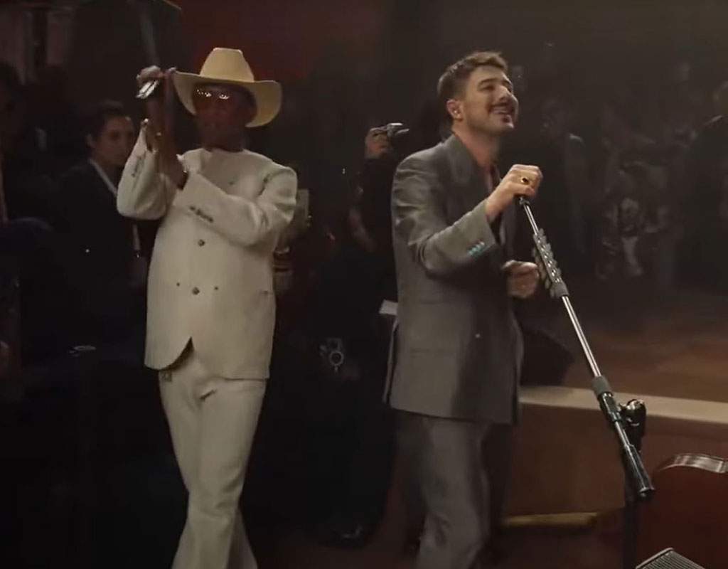 Two singers peform together. One in a white suit and stetson claps their hands. The other tilts the mic stand.