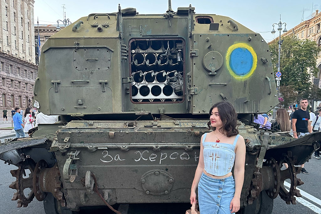 A woman stands at the back of an armoured military vehicle, the door of which is open.