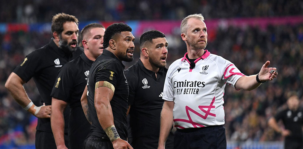 Four rugby players stand and watch beside a referee gesturing with his arm.