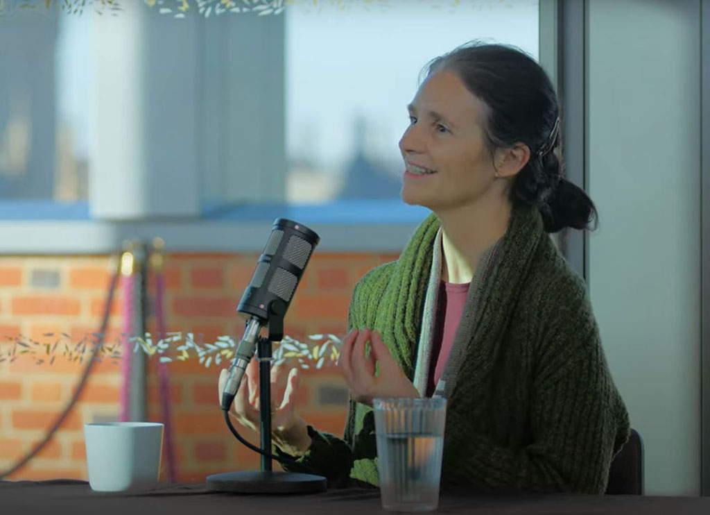 a woman sits at a table, with a mic in front of her, talking and holding her hands out in front of her.