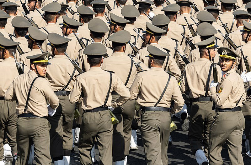 Rows of soldiers march away from the camera, two in the back row turn their heads back.