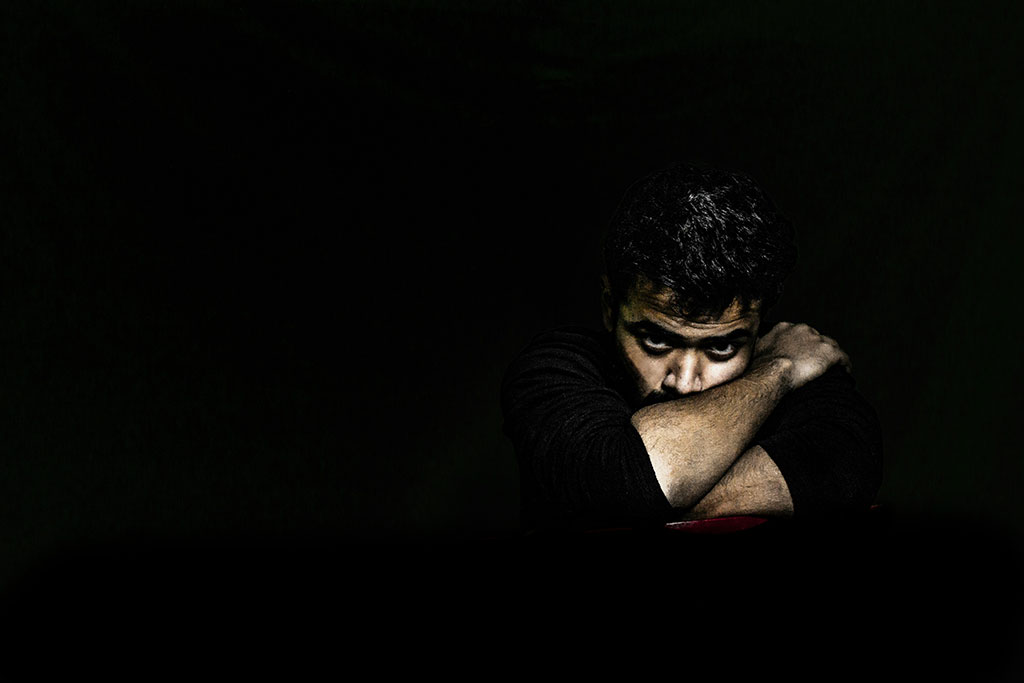 In a darkened room, a man's angry face is lit as he rests on arms folded tightly around it.