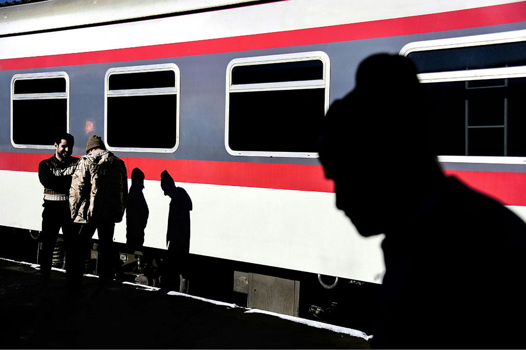 On a sun-lit railway platform, two men talk casting shadows on a waiting red and white train. Another shadow beside them is that of a man silouheted in the foreground