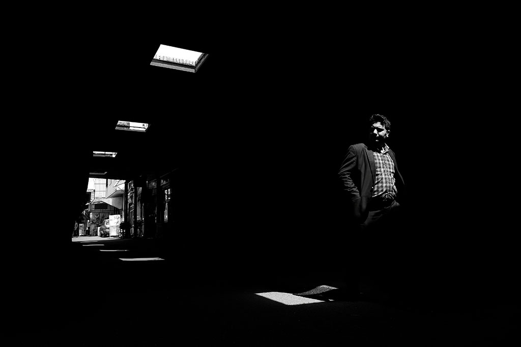 A man walks through a dark alley, looking to one side, illuminated only by roof lights.