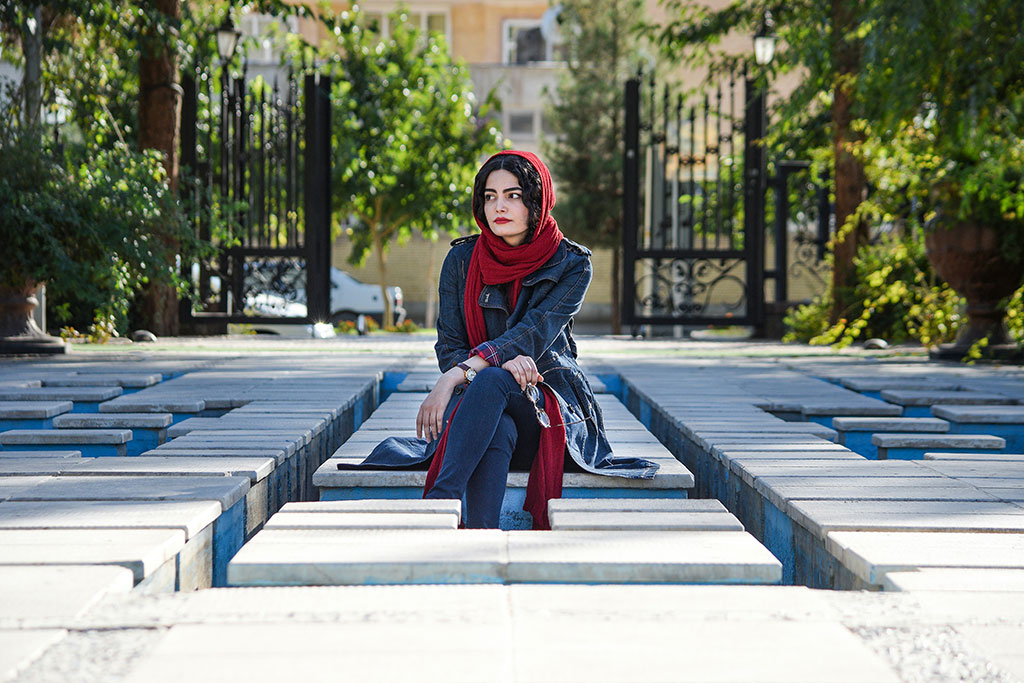 A woman wearing a headscarf sits, looking pensive, amid a grid of concrete seats.