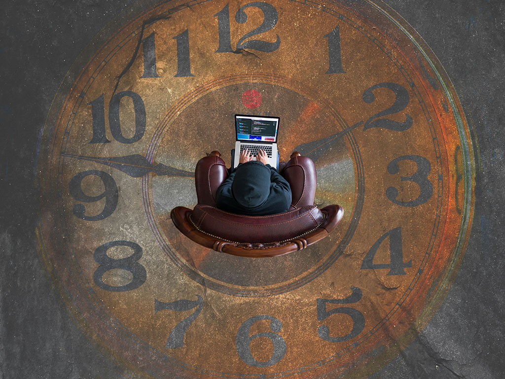 Looking straight down on someone sitting in an armchair working on a laptop. They are surrounded by clock numerals and hands on the floor.