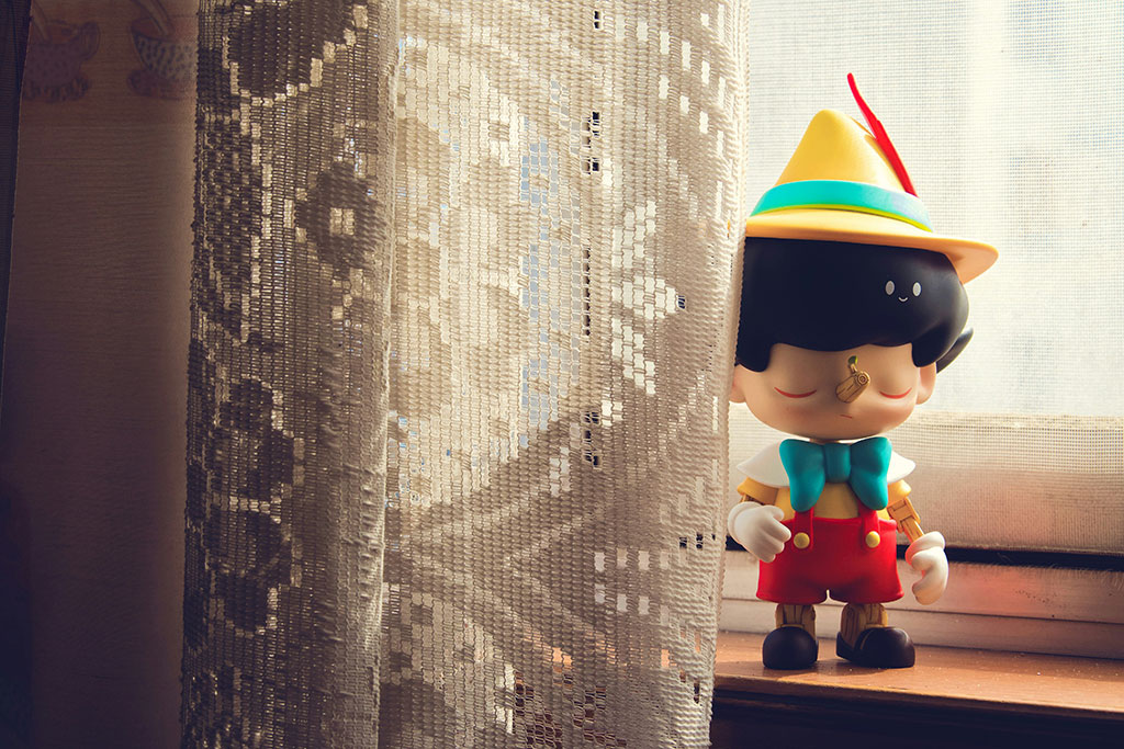 A Pinnochio figure stands on a window sill beside some net curtains.