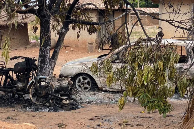 A burnt out motor cycle and car stand amid charred debris in a dusty compound.