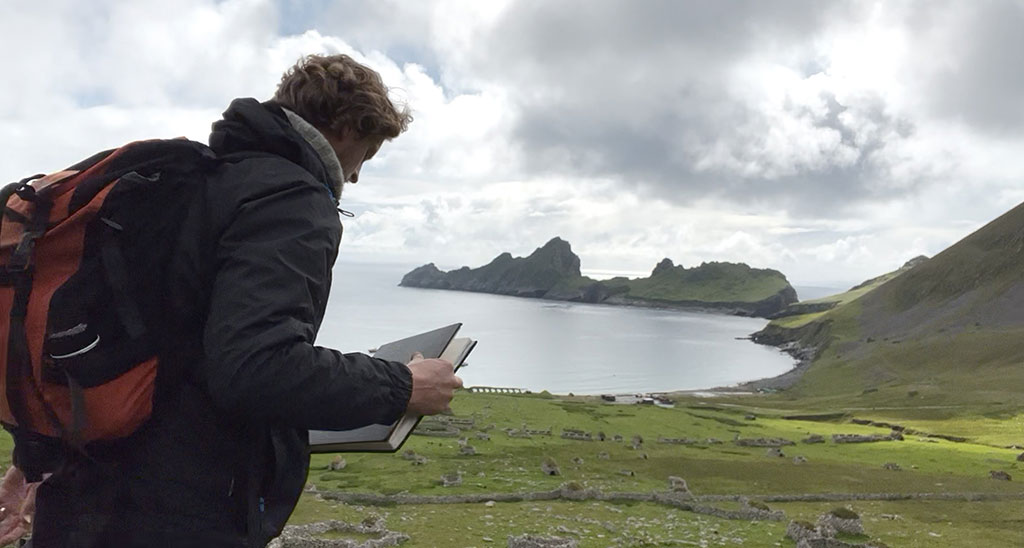 An artist holds a sketchbook while standing overlooking a deserted village by a bay, sided by jagged cliffs.