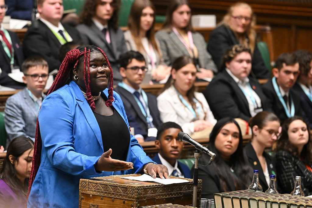 A young woman in a blue suit stands at a wooden box in a parliamentary debating chamber looking upward while speaking. 