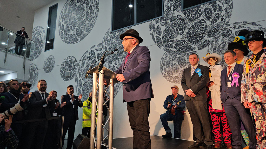 A winning candidate at an election address the audience from a lectern while the loosing candidates look on