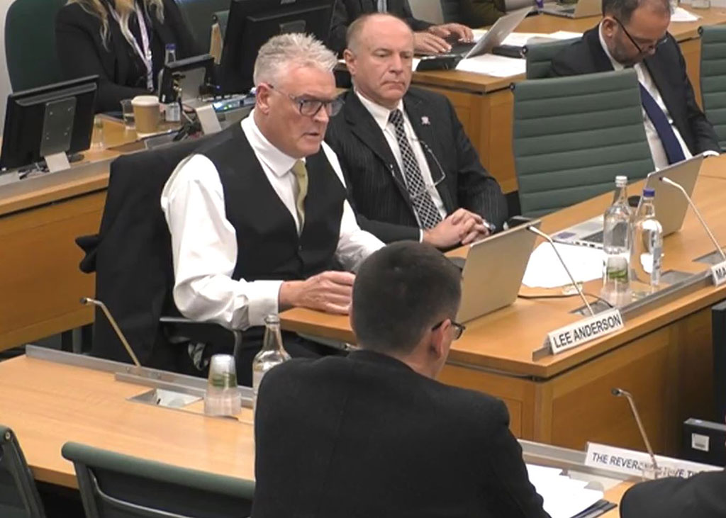 A man wearing a waist coats sits at a desk and ask a questions of a panel of people with their backs facing us.