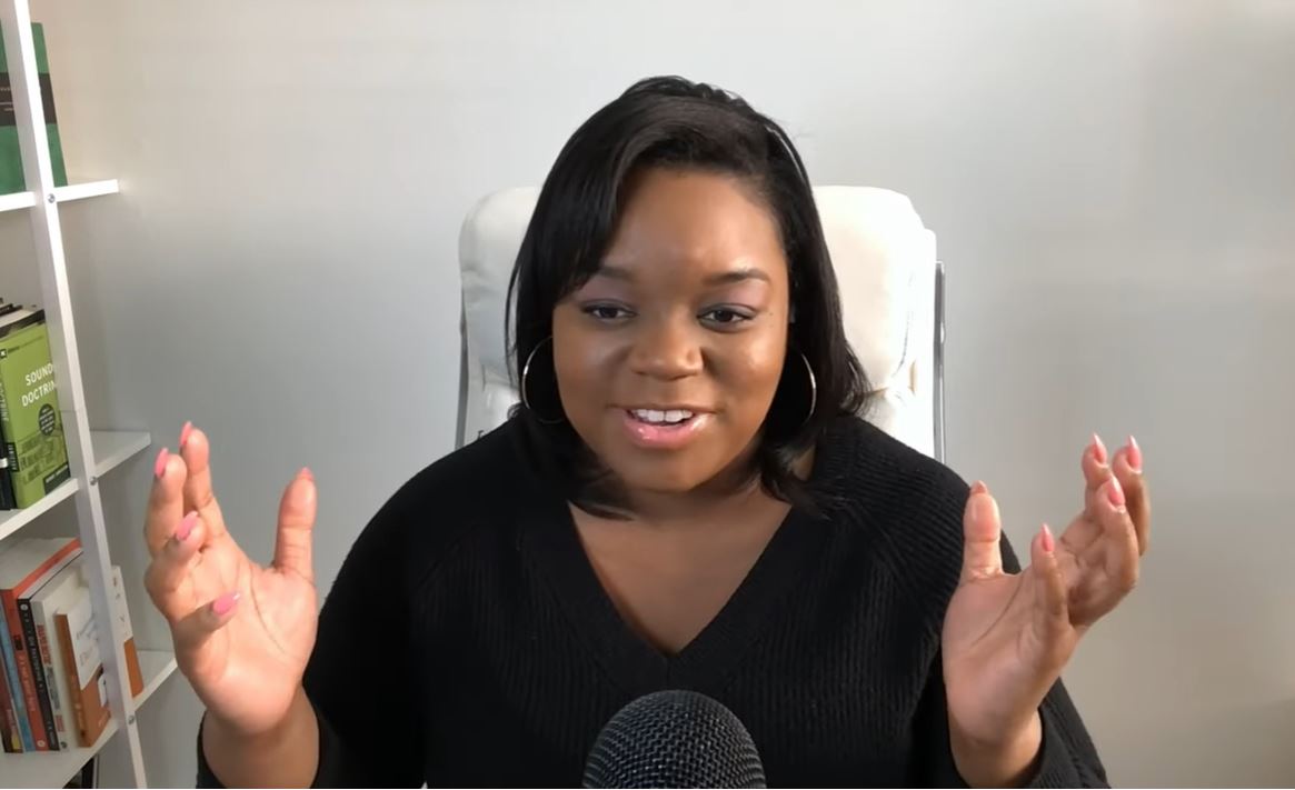 A woman sits and speaks into a desktop microphone and holds both hands up and out expressively.