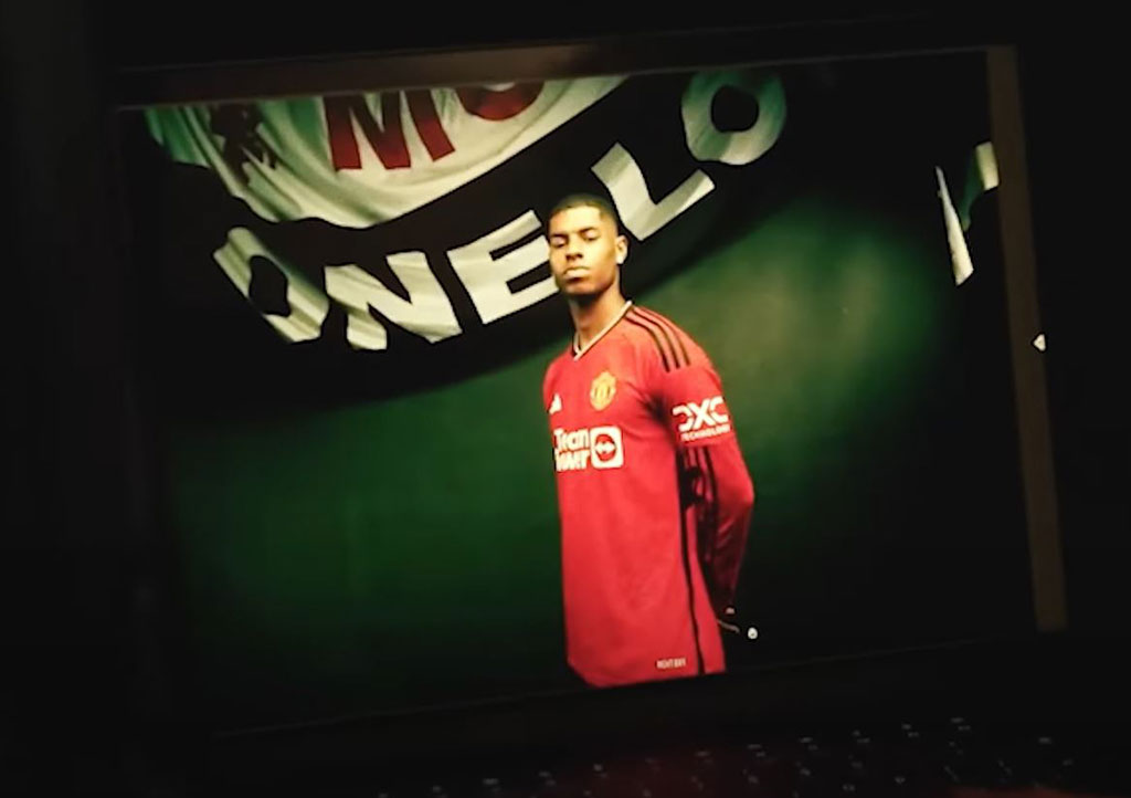 A professional footballer wearign a red top stands proudly beneath a white and black banner.