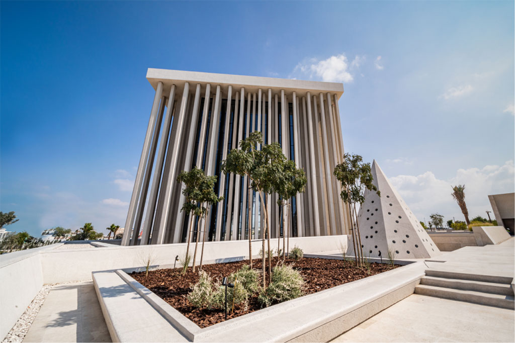 A large, modern cubiod building with a series of thin external pillars is seen from low down against a sunny blue sky 