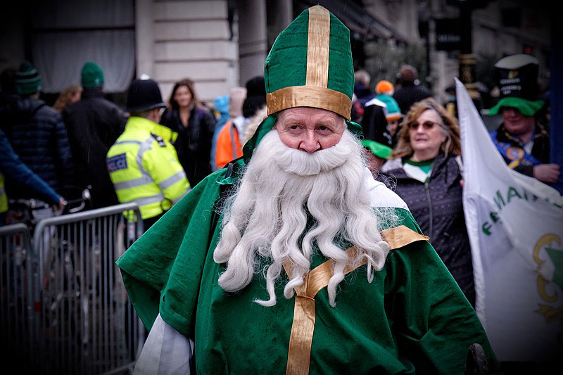 A parade particpant dressed as a bishop in green vestments with a false beard walks down a street.