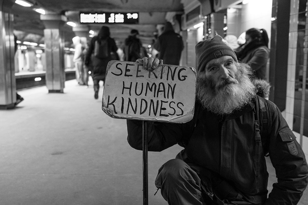 a man in a wheelchair sits in a subway station holding a sign reading 'seeking human kindness'.