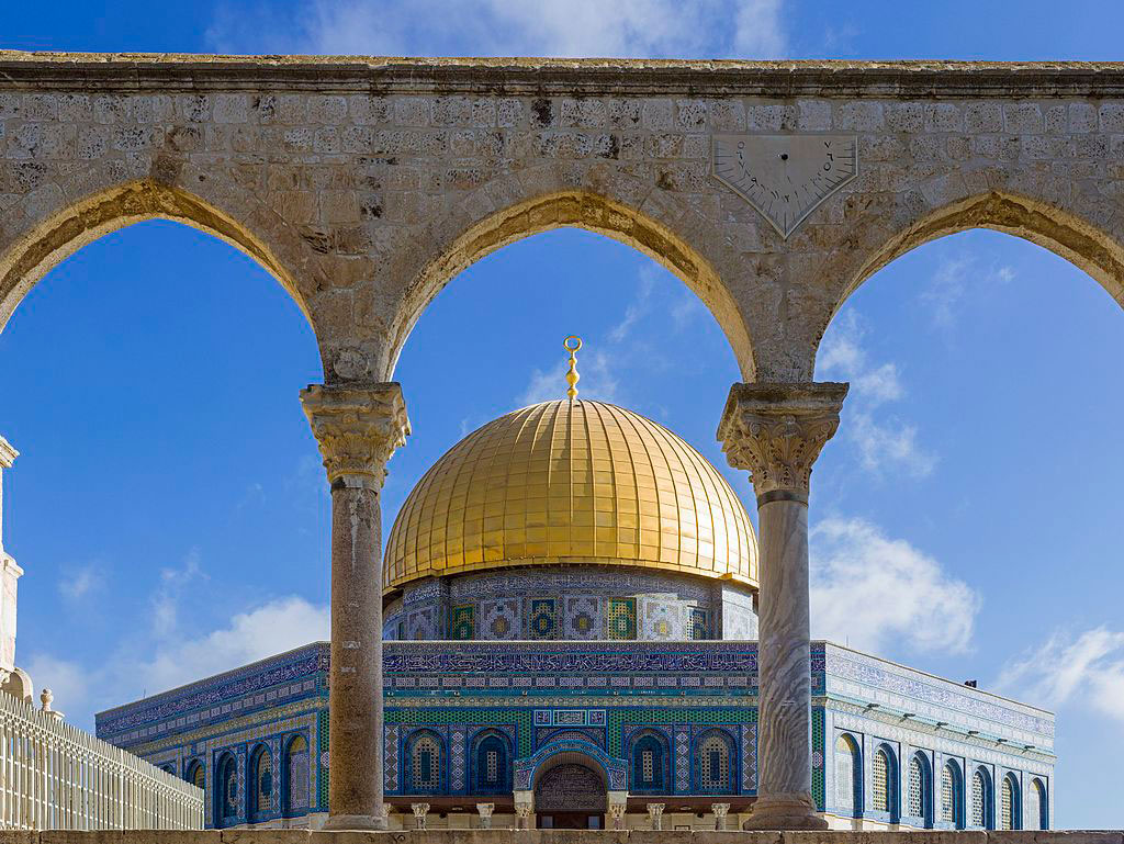 A gold-domed, blue-walled octagonal mosque seen through a row of arches.