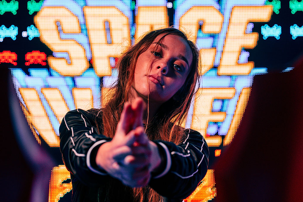 A woman stand in front of a large video screen displaying the Space Invaders title, hold her hands out in front of her.