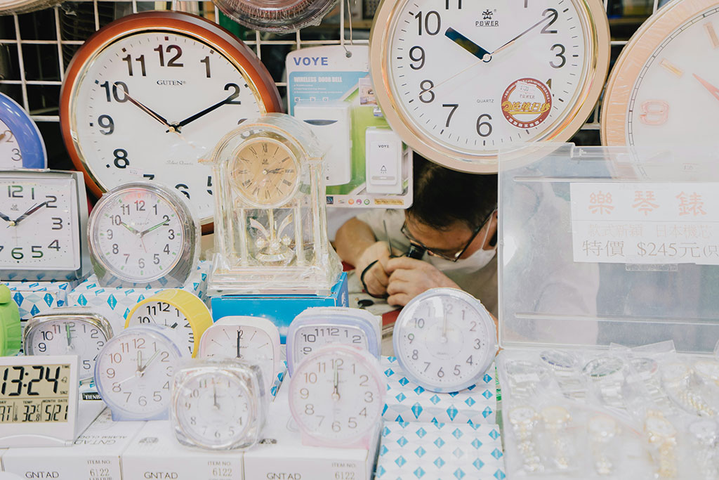 A clock repair peers at a clock he is repairing, amid a see of alarm and wall clocks on display