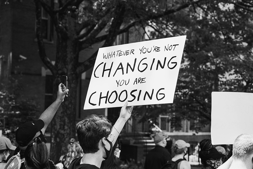 A protest placard is held above a march, reading 'If you don't change it, you choose it'.