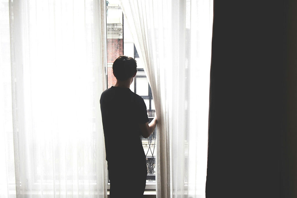 A persons stands, holding a net curtain aside to gaze out.