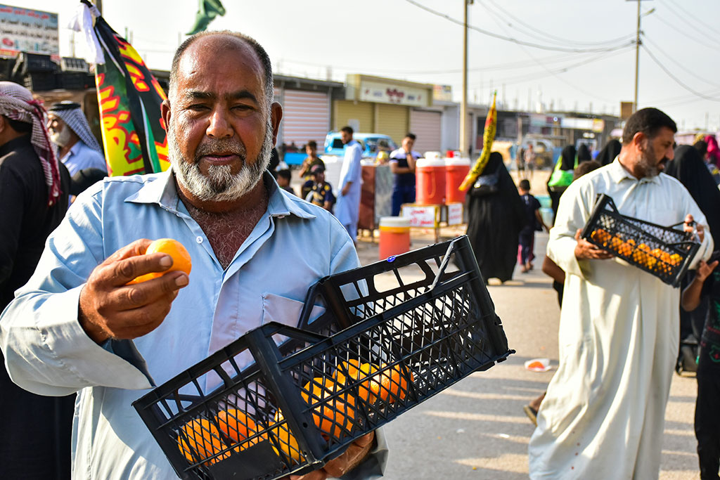 A man holds a basket of oranges and offers one to pilgrims passing by