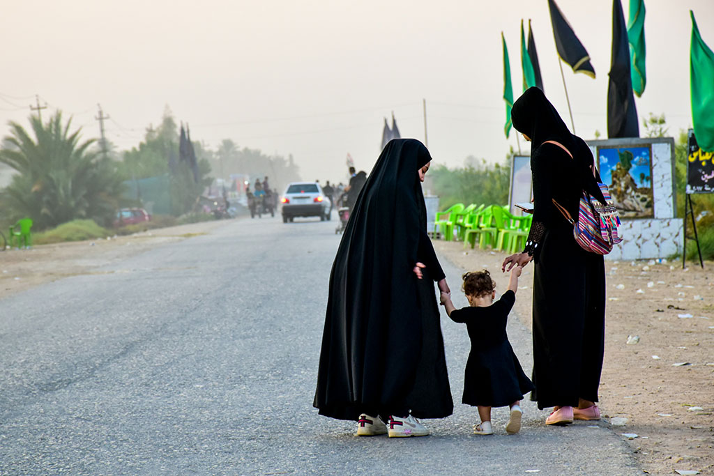 Two black clad women pligrims hold the hands of a toddler walking along the side of a road.