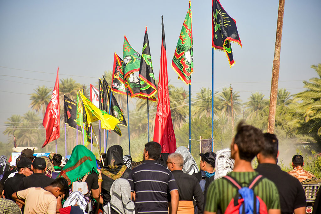 Pilgrims walk along a dusty road carrying colour flags with text on them on long poles