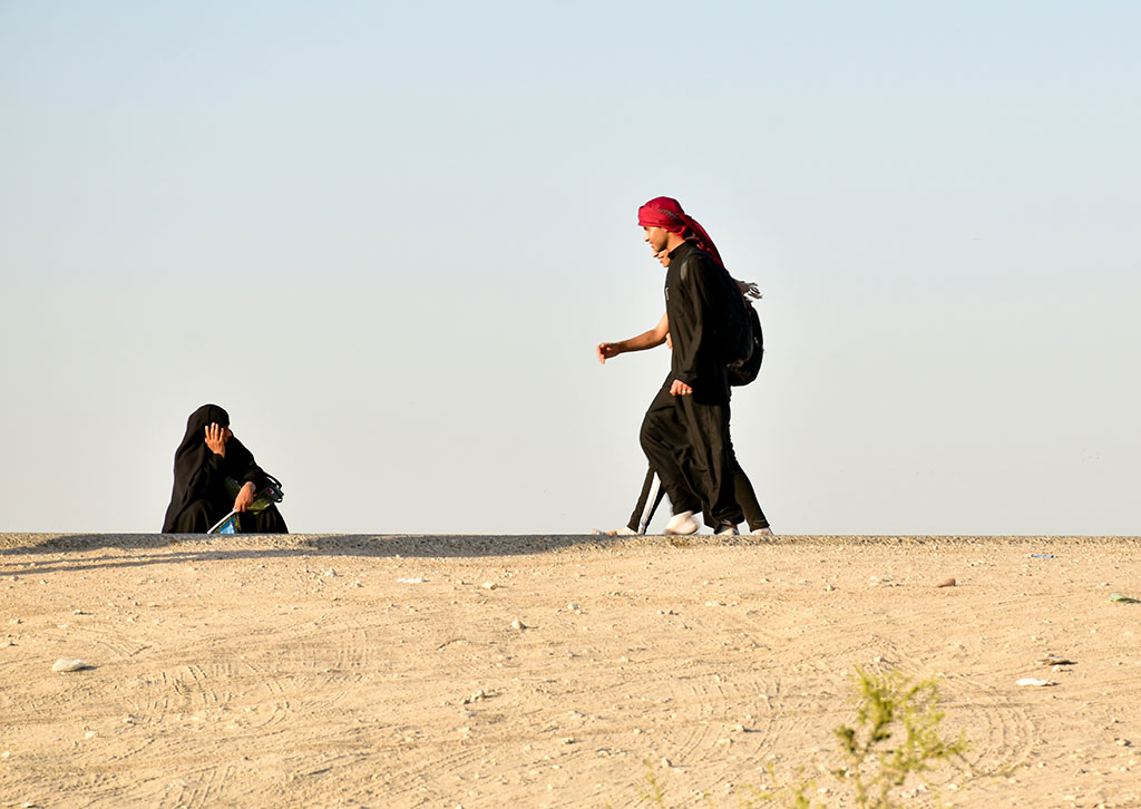 A black clad pilgrim squats and holds his head on the side of a desert road as other pilgrims walk along.